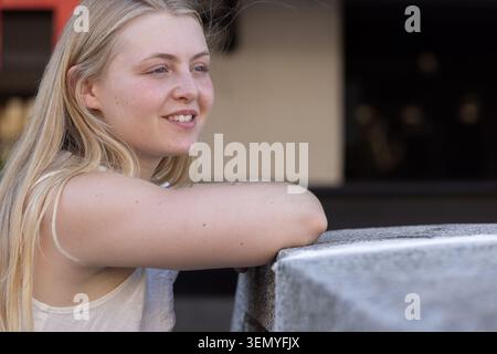Female leaning on stone ledge at urban plaza, resting arms, wearing pale sleeveless top, smiling Stock Photo