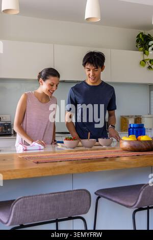 Diverse couple arranging three small bowls with red berries on wooden kitchen island, using spoon Stock Photo
