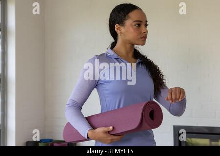 African American woman holding rolled purple mat wearing light purple zip-up top at studio window Stock Photo