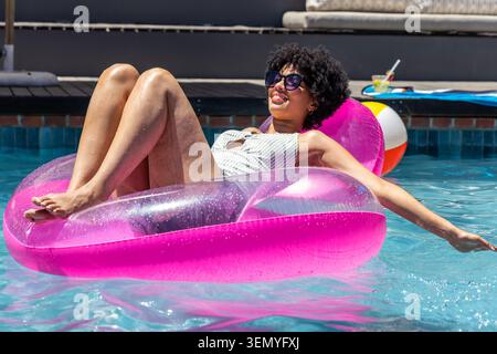 African American woman floating on pink float in pool, wearing suit, sunglasses, beach ball nearby Stock Photo