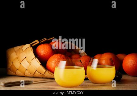 Fresh orange juice served in two glasses with ripe oranges in wicker basket on wooden table against black background. Healthy citrus drink with copy s Stock Photo