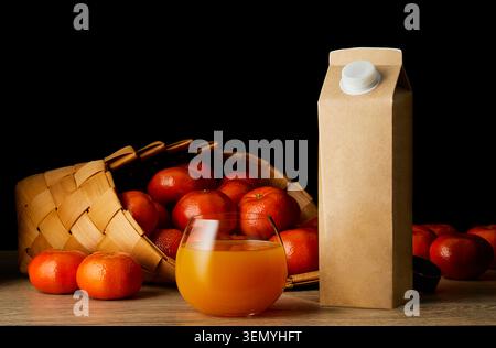 Glass of mandarin juice with kraft paper carton and fresh mandarins in wicker basket on dark background. Healthy citrus drink. Stock Photo
