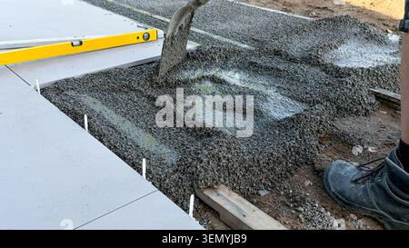 Construction worker pouring concrete mix into a mold on a building site, with leveling tool and partially completed pavement visible in the background Stock Photo