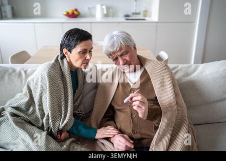 Old covered in blankets checking temperature level with thermometer sitting on sofa at home Stock Photo