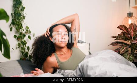 Relaxed Woman Waking Up in Cozy Bedroom With Plants, Soft Morning Light and Tattoos Stock Photo