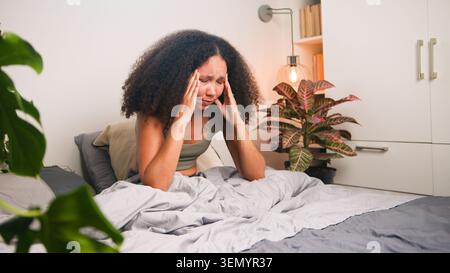 Black Woman Sitting in Bed Holding Head in Pain With Headache and Morning Stress in Bedroom Stock Photo