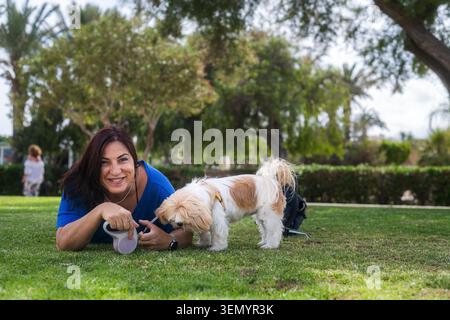 A smiling woman with her Shih Tzu lying on the grass. Outdoor pet lifestyle. She smiles affectionately at her dog. A small, brown and white dog sniffs Stock Photo