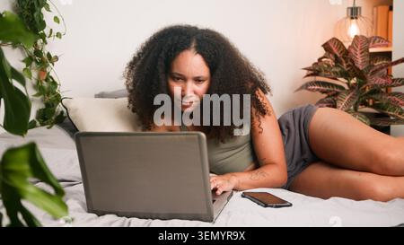 Woman Working on Laptop in Cozy Bedroom Surrounded by Plants and Smartphone Stock Photo