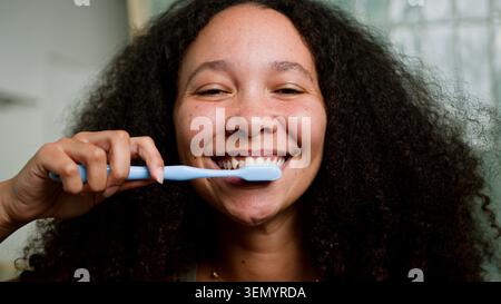 Young Woman Smiling While Brushing Teeth With Blue Toothbrush In Morning Bathroom Routine Stock Photo