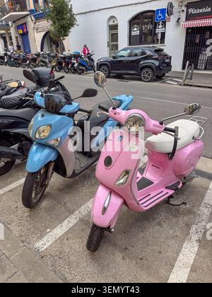 Sitges, Spain -  October 02, 2025: A high-angle shot focusing on a pastel pink vintage-style Vespa scooter and a light blue modern moped parked next t Stock Photo