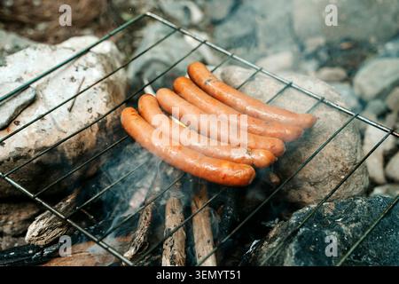 Sausages sizzling on a metal grill grate over an open fire, surrounded by rocks and wood, capturing the essence of outdoor camping and rustic cooking. Stock Photo