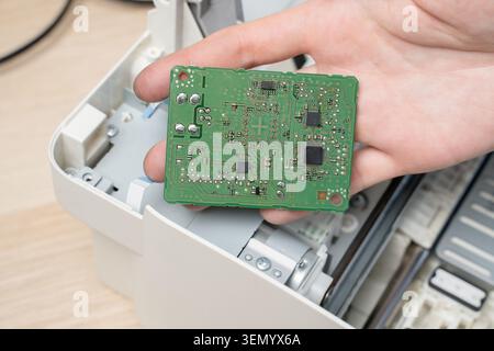 A technician's hand holds circuit board removed from a disassembled printer, revealing chips, solder points and traces during electronic repair, diagn Stock Photo