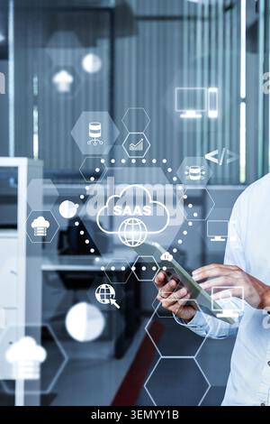 User interacts with mobile device while monitoring SaaS on screen in office setting Stock Photo