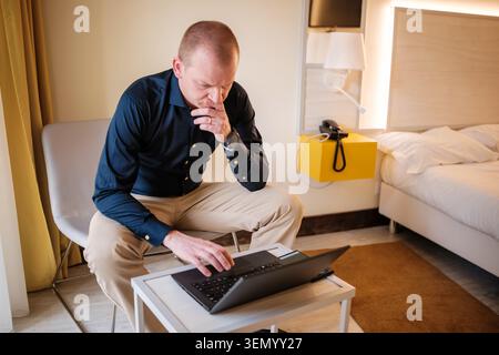 Thoughtful businessman using laptop, working remotely from a hotel room Stock Photo