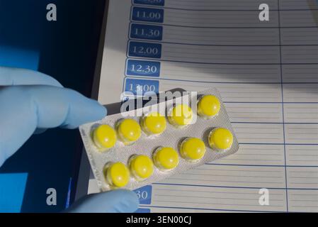 Hand in a blue medical glove holding a blister of bright yellow pills in sunlight over a page from a daily planner, close-up. Stock Photo