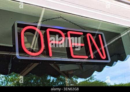 A vibrant red neon sign spelling out the word OPEN hangs in a glass window, reflecting the sky and trees outside. Stock Photo