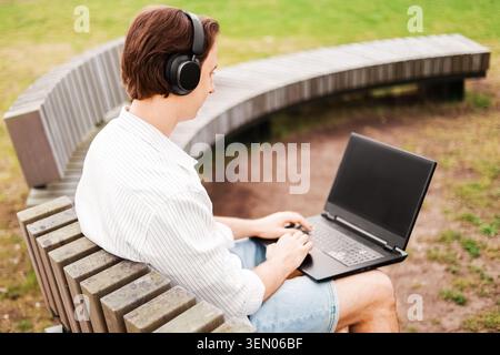 Man doing remote freelance work on a laptop while wearing headphones on a park bench outdoors. High quality photo Stock Photo