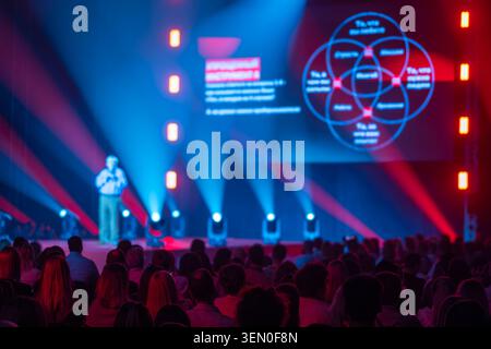 Conference speaker presents on stage to attentive audience, colorful lights and projected slides create dynamic, professional atmosphere. Stock Photo
