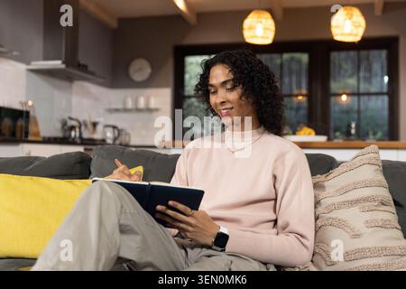 Non-binary person writing in dark-blue notebook and pen on gray sofa at home, wearing pink sweater Stock Photo