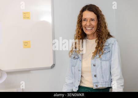 Mature woman laughing in office by whiteboard with 2 yellow sticky notes, denim jacket, copy space Stock Photo