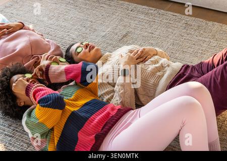Three women lying on woven rug in living room, wearing knit sweaters, holding cucumber slices Stock Photo