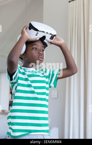 African American boy holding white VR headset up at home wearing white headphones and green shirt Stock Photo