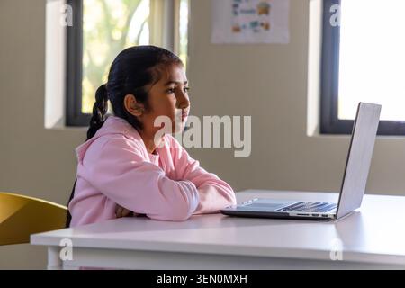Asian school-age girl studying at classroom desk, using open laptop while wearing pink hoodie Stock Photo
