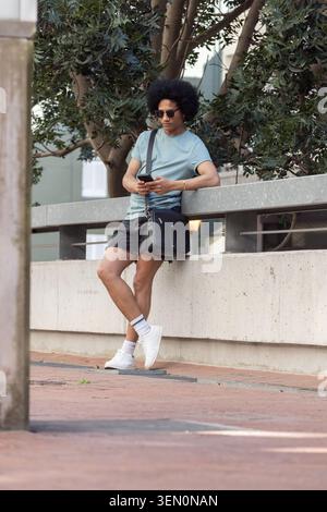 African American man leaning on concrete railing wearing sunglasses on walkway, checking smartphone Stock Photo