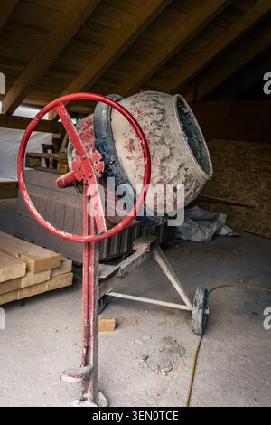 A close-up shot of a red and gray concrete mixer standing on wheels within an unfinished construction site. The mixer is covered in remnants of cement Stock Photo