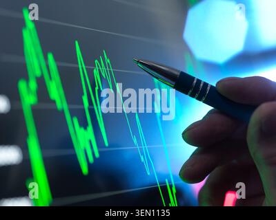 Hand holding a pen pointing at a fluctuating green line graph on a screen. Stock Photo