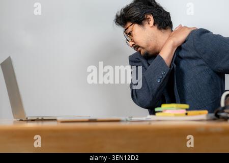 A young businessman suffering from neck and shoulder pain while working at his desk in the office, depicting symptoms of office syndrome. Stock Photo