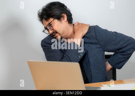 A young businessman suffering from neck and shoulder pain while working at his desk in the office, depicting symptoms of office syndrome. Stock Photo