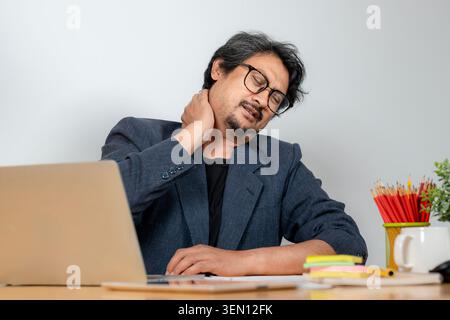A young businessman suffering from neck and shoulder pain while working at his desk in the office, depicting symptoms of office syndrome. Stock Photo