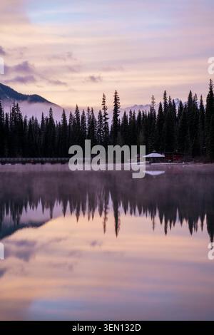 Tranquil reflection of pine trees and mountains in a serene lake at sunrise with misty skies. Stock Photo