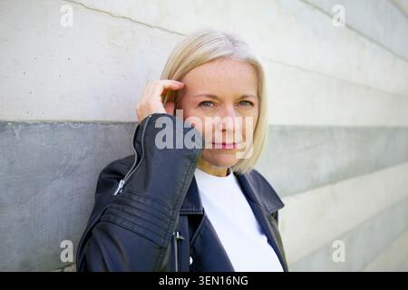 Mature woman in black leather jacket leaning against a modern concrete wall. Stock Photo