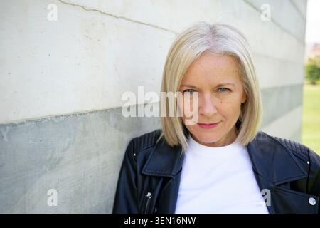 Blonde woman in a leather jacket leans against a white and gray concrete wall outside. Stock Photo