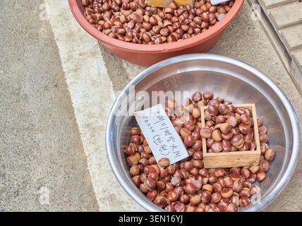 Raw brown tree nuts in bowls with traditional wooden measuring cups and Korean price signs at a local outdoor street bazaar in South Korea. Stock Photo