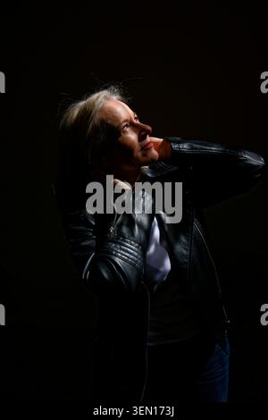 Woman in leather jacket looks upwards illuminated against a dark background. Stock Photo