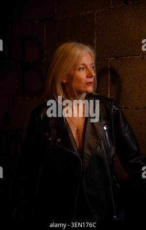 Woman in leather jacket stands thoughtfully against a shadowy brick wall with light on her face. Stock Photo