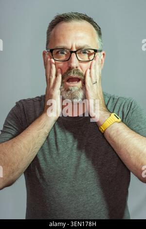 A man stands in a studio with a shocked expression. He has gray hair and glasses. His hands are on his face as if reacting to unexpected news or a sur Stock Photo