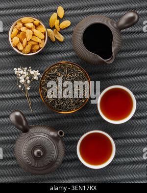 Top view of Chinese black tea set with loose leaf tea, teapot, pitcher, teacups and golden raisins on dark woven background. Stock Photo