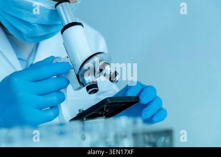 Close-up of a masked researcher examining samples through a microscope in a clean lab environment, suitable for medical research, diagnostics, biotech Stock Photo