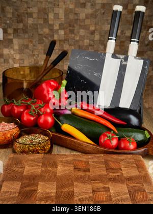 Wooden end grain cutting board with fresh vegetables, chef knives, seasonings and kitchen utensils on wooden table. Stock Photo