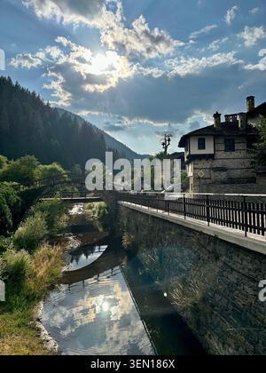 Sunlight breaks through mountain clouds above a mirror-like village canal, turning the valley into liquid gold. Stock Photo