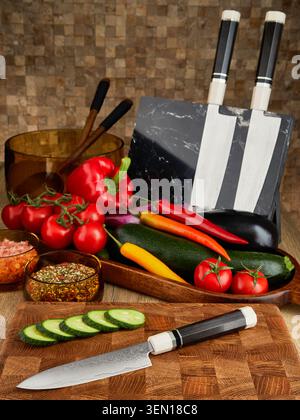 Chef knife on wooden cutting board with sliced cucumber, fresh vegetables, spice bowls and kitchen utensils on wooden table. Stock Photo