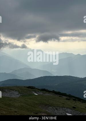 Dark storm clouds press over layered mountain ridges while pale shafts of rain and light sweep across the distant valley. 3 Stock Photo