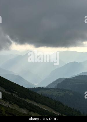 Dark storm clouds press over layered mountain ridges while pale shafts of rain and light sweep across the distant valley. 2 Stock Photo