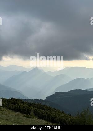 Dark storm clouds press over layered mountain ridges while pale shafts of rain and light sweep across the distant valley. 1 Stock Photo