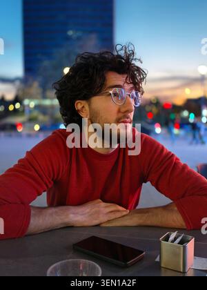 Thoughtful Young Man Wearing Glasses and Red Sweater Sitting at Outdoor Table in City Evening Stock Photo