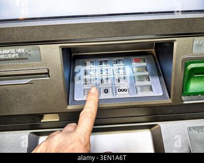 A hand is pressing a button on an ATM keypad, indicating an ongoing transaction for cash withdrawal in a public area. Stock Photo